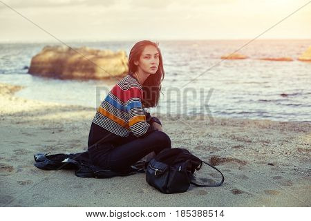 A girl sitting on the sand beach by the sea with beautiful sunset on the background. Woman in jeans pullover and jacket on the sand