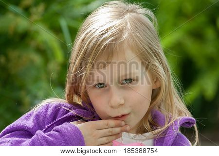 a portrait of a little girl in a lilac jacket on the background of green leaves in the garden, a hand near the mouth, a little gossip, a secret to tell sarcastically, wheat hair,portrait in front