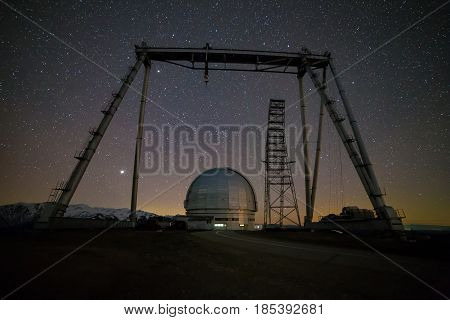 Night view. A special astrophysical observatory and a crane against the background of the starry sky and snowy peaks of the Arkhyz mountains.