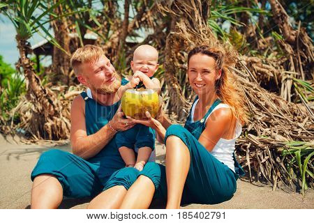 Happy family funny picnic on beach - mother father feed baby boy. Little son eat fruits with fun and drink fresh coconut. Healthy lifestyle people walking activity on summer vacation with child.