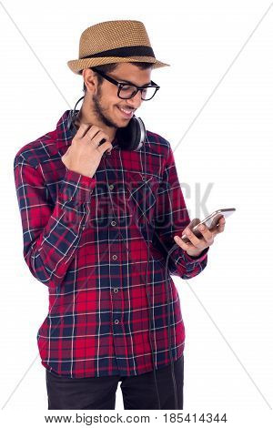 Happy friendly young man using mobile phone by his hand and holding a headphones by the other hand guy wearing red caro shirt and jeans with glasses and beige hat isolated on white background