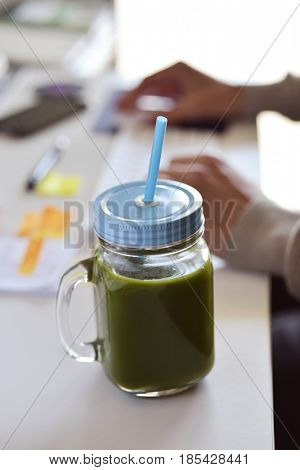 closeup of a green detox smoothie served in a glass jar on an office desk, and a young caucasian man using his computer in the background