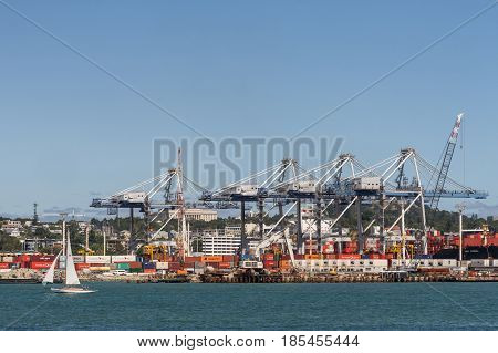 Auckland New Zealand - March 3 2017: The container terminal with its cranes and boxes under blue sky and behind greenish ocean water. White sailing boat in front. War Memorial Museum in back.
