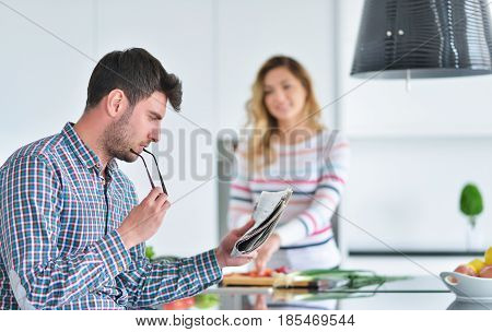 Couple standing and sitting at the kitchen while smiling and man reading a newspaper and holding mug before work