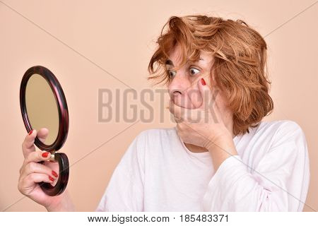 Shocked and surprised middle aged woman looking at her skin and unkempt hair in the mirror