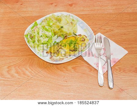 Pork chop baked with the onion mushrooms and cheese with the garnish of mashed potatoes and salad of fresh young cabbage on the dish fork and knife on the paper napkin beside on a wooden surface