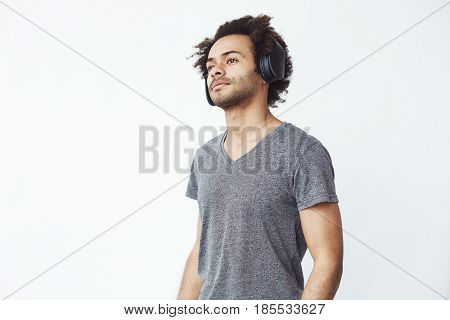 Young handsome african man listening to streaming music in black over-the-ear wireless headphones over white wall, noding to beats. Copy space.