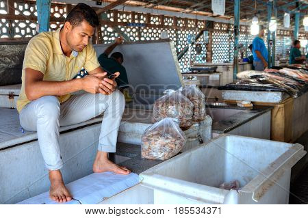 Bandar Abbas Hormozgan Province Iran - 16 april 2017: One young Iranian man sitting at the entrance to the fish market near the packages with shrimp and reads message on the smartphone screen.