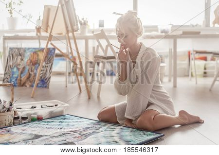 Beautiful young woman sitting on the floor in a bright Studio and paints with paint on canvas.
