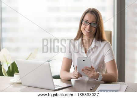 Portrait of happy businesswoman at desk holding tablet in hands, looking at camera. Female entrepreneur using phablet in office. Smiling woman inspects electronic documentation, digital office work