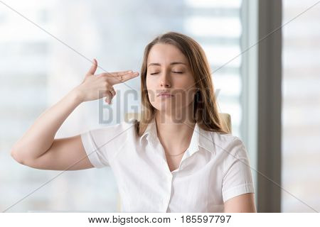 Woman with indifferent facial expression puts a gun composed from fingers to temple. Female employee showing complete apathy to work. Figuratively suicide because of chronic fatigue or stress at work