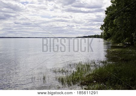 Wide landscape view of Lake St-Louis in Notre Dame de Ile Perrot, Quebec with trees and foliage on the right side of the frame on a sunny day in June.