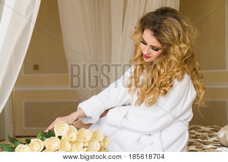 Beautiful seductive woman flirting with the camera is sitting on the bed with a big bouquet of white roses in White bathrobe looking up with a coquettish look.