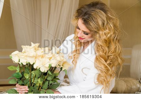 Beautiful seductive woman flirting with the camera is sitting on the bed with a big bouquet of white roses in White bathrobe looking up with a coquettish look.