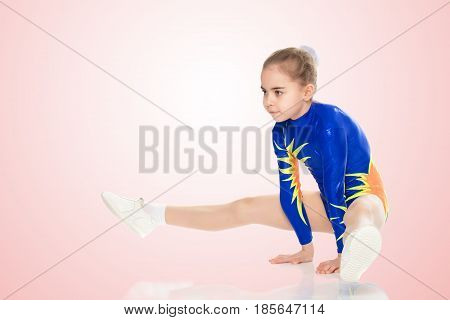 Beautiful Russian girl gymnast younger school age, in blue in a sports swimsuit.The girl performs a difficult acrobatic exercise.Pale pink gradient background.