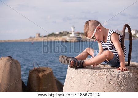 beautiful little boy in sunglasses sits on a concrete breakwater against the background of the sea and the shore with lighthouse