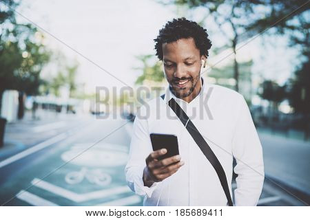 Happy american African man using smartphone outdoor.Portrait of young black cheerful man texting a sms message while listining to music.Blurred background, horizontal