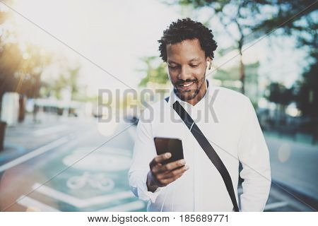 Happy american African man using smartphone outdoor.Portrait of young black cheerful man texting a sms message while listining to music.Blurred background, horizontal.Flare effects