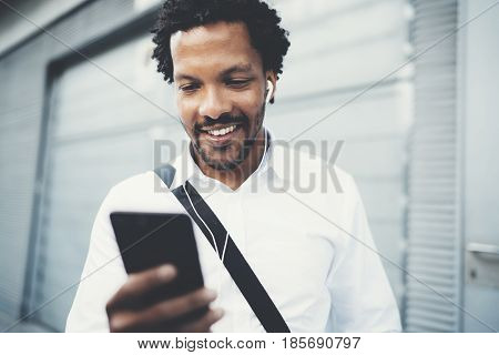 Portrait of Smiling African American man in headphones standidng in sunny street listening to songs on his mobile phone, using Internet-enabled electronic device, texting friends.Blurred background