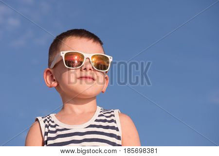 smiling little boy wearing white sunglasses with sea sunset reflection and sailor stripes shirt on blue sky background with copyspace