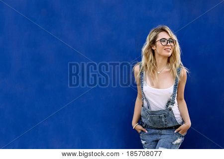 Young Stylish Blond Girl In Denim Overalls Made Of Denim And On A Blue Background