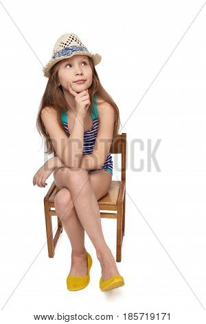 Full length child girl in swimsuit and summer hat sitting on wooden chair thinking contemplating looking up at blank copy space, studio portrait