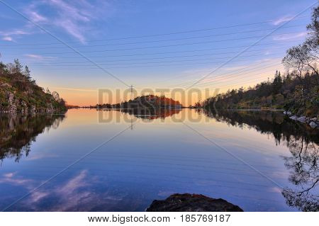 Colorful sunset with sky reflections in lake. Power lines and dam in background. Shot in Norway Europe.