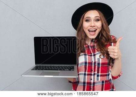 Portrait of a smiling happy girl in plaid shirt holding blank screen laptop computer and showing thumbs up gesture isolated over gray background