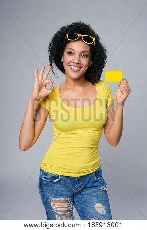 Smiling mixed race african american caucasian woman holding credit card and gesturing OK, isolated on grey background
