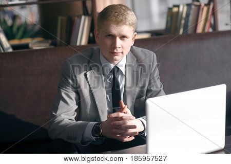 young businessman discussing business sitting at the table. looking right at you, folding his fingers together. next on the table is a laptop