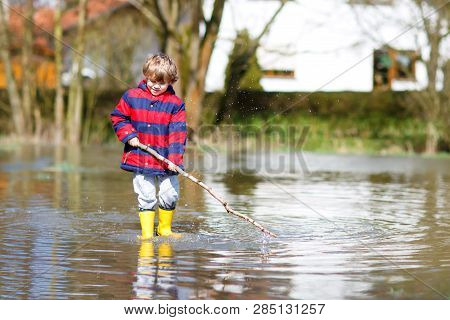 Happy Little Kid Boy In Yellow Rain Boots Playing With Paper Ship Boat By Huge Puddle On Spring Or A