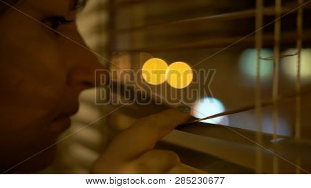 Young Brunette With Brown Eyes Peeks Through The Window With Her Finger, Pushing Open The Blinds
