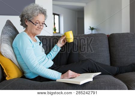 Happy Positive Lady Enjoying Crime Novel. Retired Senior Woman In Casual And Eyeglasses Drinking Tea