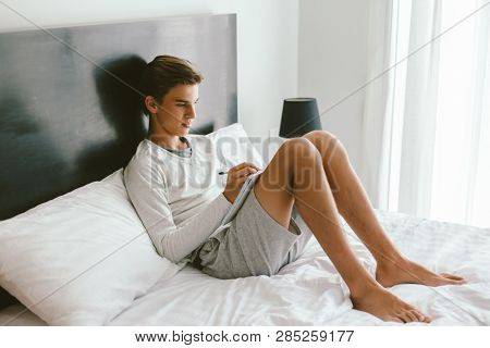 16 years old teenage boy writing school notes or doing homework while relaxing on couch in his room