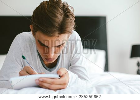 16 years old teenage boy writing school notes or doing homework while relaxing on couch in his room