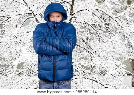 Sad Asian man in blue down padded coat, against snowy winter background. Man outdoors in warm winter clothes. Snow and cold