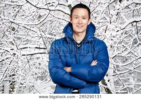 Happy Asian man in blue down padded coat, against snowy winter background. Man outdoors in warm winter clothes. Snow and cold