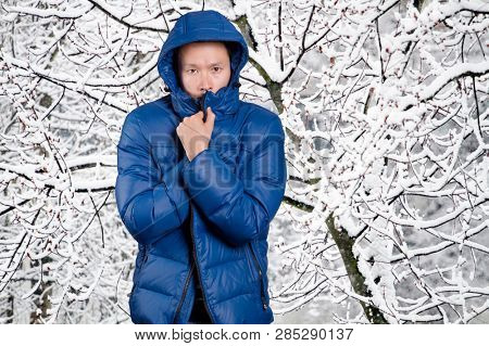 Sad Asian man in blue down padded coat, against snowy winter background. Man outdoors in warm winter clothes. Snow and cold
