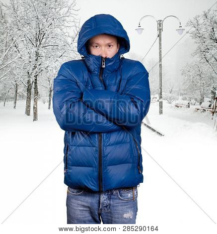Sad Asian man in blue down padded coat, against snowy winter background. Man outdoors in warm winter clothes. Snow and cold