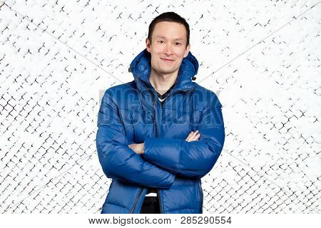 Happy Asian man in blue down padded coat, against snowy winter background. Man outdoors in warm winter clothes. Snow and cold