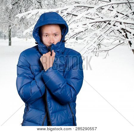 Sad Asian man in blue down padded coat, against snowy winter background. Man outdoors in warm winter clothes. Snow and cold
