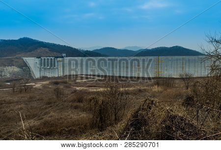 Winter Landscape Of Field Of Dry Grass And Trees In Front Of Concrete Dam  With Yellow And Black Wat