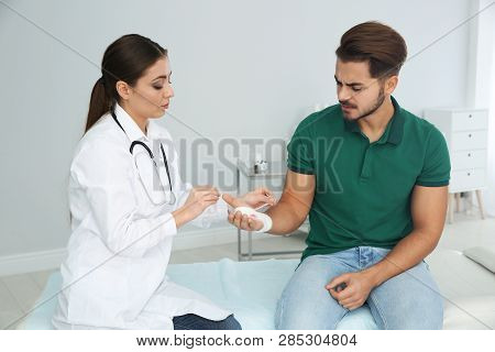 Female Doctor Applying Bandage On Young Man's Hand In Clinic. First Aid