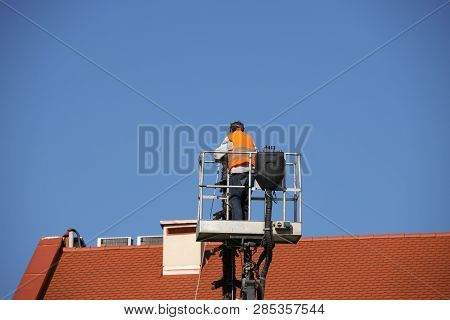 The Worker In Overalls Works At Height In A Building Mechanical Lifting Basket. Repair And Construct