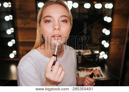 Young Blonde Woman Applying Lipgloss On Lips. She Look On Camera. Serious And Concentrated. Mirror W