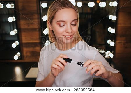 Cheerful Nice Young Woman Hold Lipgloss And Look On It. She Stand In Beauty Room. Mirror With Light 
