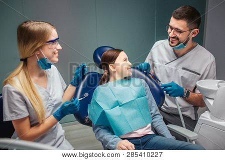 Positive Happy Girl Sit In Dental Chair And Look And Male Dentist. He Smile To Her. Female Helper Ha