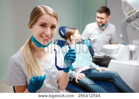 Young Female Dentist Look On Camera And Smile. She Hold Glasses. Male Dentist Stand Beside Girl. Chi