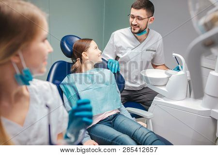 Child Sit In Denta Lchair In Room. Male Dentist Stand Beside Her And Smile. Young Woman Look At Them