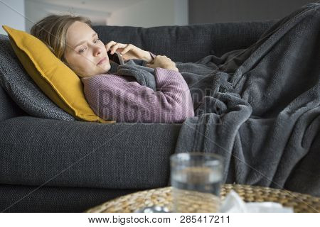 Sick Young Caucasian Woman In Purple Sweater Covered With Grey Blanket Lying On Grey Sofa In Living 